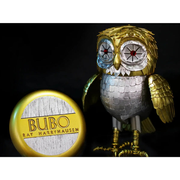 A collectible Bubo owl figurine by Ray Harryhausen, featuring a metallic gold and silver body with blue-tinted eyes, shown next to its circular gold and silver Bubo collectible capsule on a black background.
