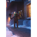 A collectible figurine of Kermit the Frog dressed in winter attire, standing on a snowy city sidewalk at night next to a brick building with a lit window.