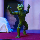 A green action figure of a monstrous creature with horns and an open mouth, posed with arms raised, standing on a red carpet in front of a dimly lit set background.