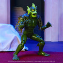 A green cartoon-style monster action figure posed in a crouching stance on a red surface, with a blurred indoor set background and visible printed copyright text at the bottom.