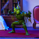 A green action figure of a monster character posed crouching on a red carpet in an indoor room setting with a bookshelf and chair in the background.