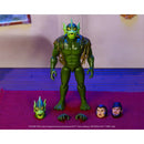A green action figure of a monster or creature stands on a purple surface, with two alternate heads and two hands placed in front. The background shows a beige wall and blue-lit floor.