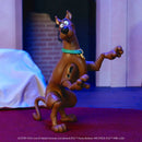 A Scooby-Doo action figure standing pose on a red rug, with a blurred indoor background and visible Scooby-Doo branding at the bottom of the image.