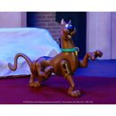 A Scooby-Doo action figure posed mid-leap on a red carpet indoors, with a white draped surface and dark background behind it.