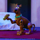 Scooby-Doo action figure posed standing on a red carpet with a sandwich in one paw, set against a dimly lit indoor background with a bed visible behind it.