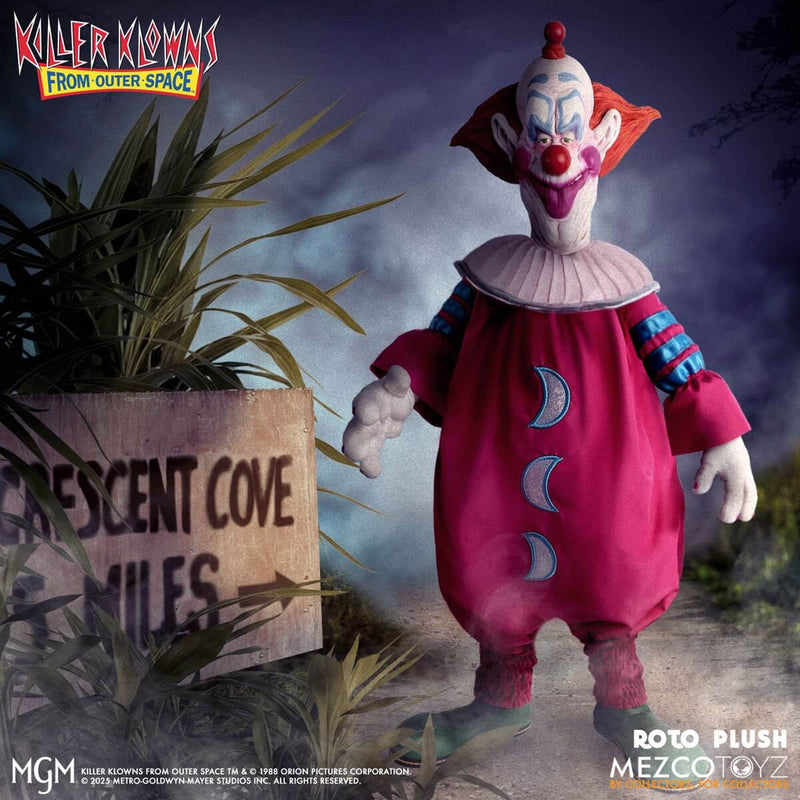Plush toy of a clown from 'Killer Klowns from Outer Space' by Mezco Toyz, standing next to a sign in a dark, eerie setting.