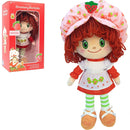 Strawberry Shortcake plush doll shown next to its red retail box on a white background. The doll has red curly hair, a pink strawberry-patterned bonnet, a white dress with red trim and green leaf embroidery, green-and-white striped tights, and tan shoes.