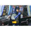 A Batman action figure posed holding a small object near its face, wearing a gray suit, dark blue cape and gloves, and a yellow utility belt, set against a blurred city background with cars.