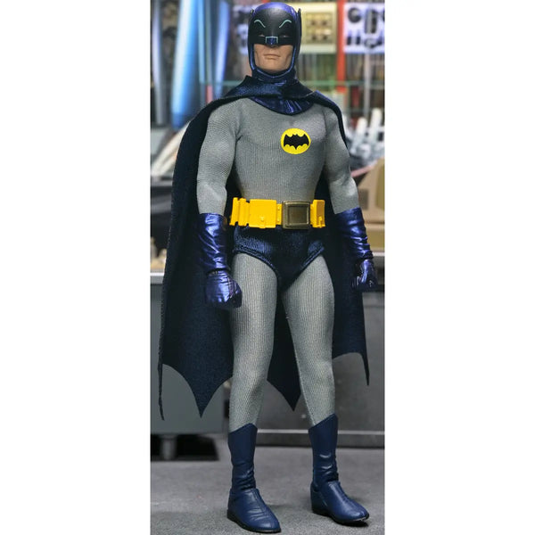 A Batman action figure or collectible doll wearing a gray textured suit, dark blue cape and boots, a yellow utility belt, and a yellow bat symbol on the chest, standing against an indoor blurred background.