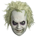 A Beetlejuice-style Halloween mask with pale off-white skin, wild blond hair, dark purple eye makeup, and green mossy accents, shown against a plain white background.