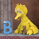 Yellow puppet with a teddy bear and a blue letter 'B' on a brick ledge.