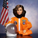 Doll in an orange astronaut suit with NASA patches, holding a helmet and gloves, against a blue sky background with an American flag.