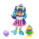 A toy doll with bright blue hair, purple polka-dot hat and bow, purple patterned sleeves, a pink skirt, green-and-white striped tights, and blue shoes, shown on a white background with a small purple-and-green owl figure and a purple hairbrush accessory.