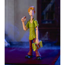 A collectible action figure of Shag from Scooby-Doo standing on a dark red rug in a dimly lit room. Shag is wearing his signature green shirt and brown pants, holding a yellow box in one hand and a small object in the other.