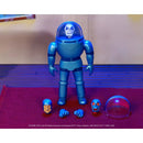 A blue action figure standing on a red surface with its accessories arranged in front, including a separate head, hands, and a clear dome. The background is an indoor setting with a beige wall and blue-lit floor.