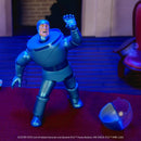 A metallic blue action figure posed mid-action on a red carpet, with a matching helmet placed beside it. The background is a dimly lit indoor set with blue lighting, including a chair and brick wall.