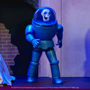 A blue metallic action figure of the Scooby-Doo WB Shield character, standing on a dark surface against a backdrop of a brick wall and a textured wall under blue lighting.